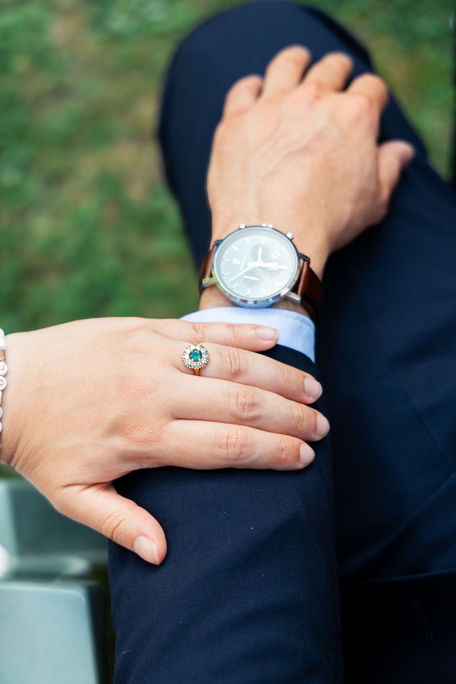 Close-up of a woman's hand with an emerald engagement ring resting on a man's arm wearing a brown leather watch.