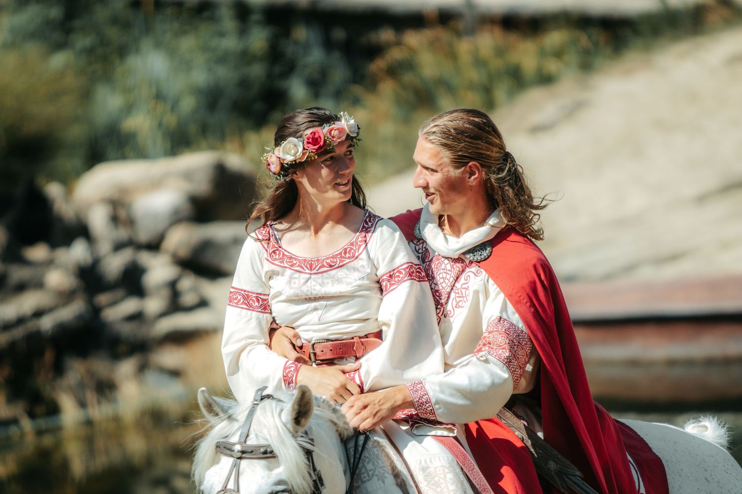 Two women in traditional Slavic folk costumes with floral wreaths smile at each other outdoors.