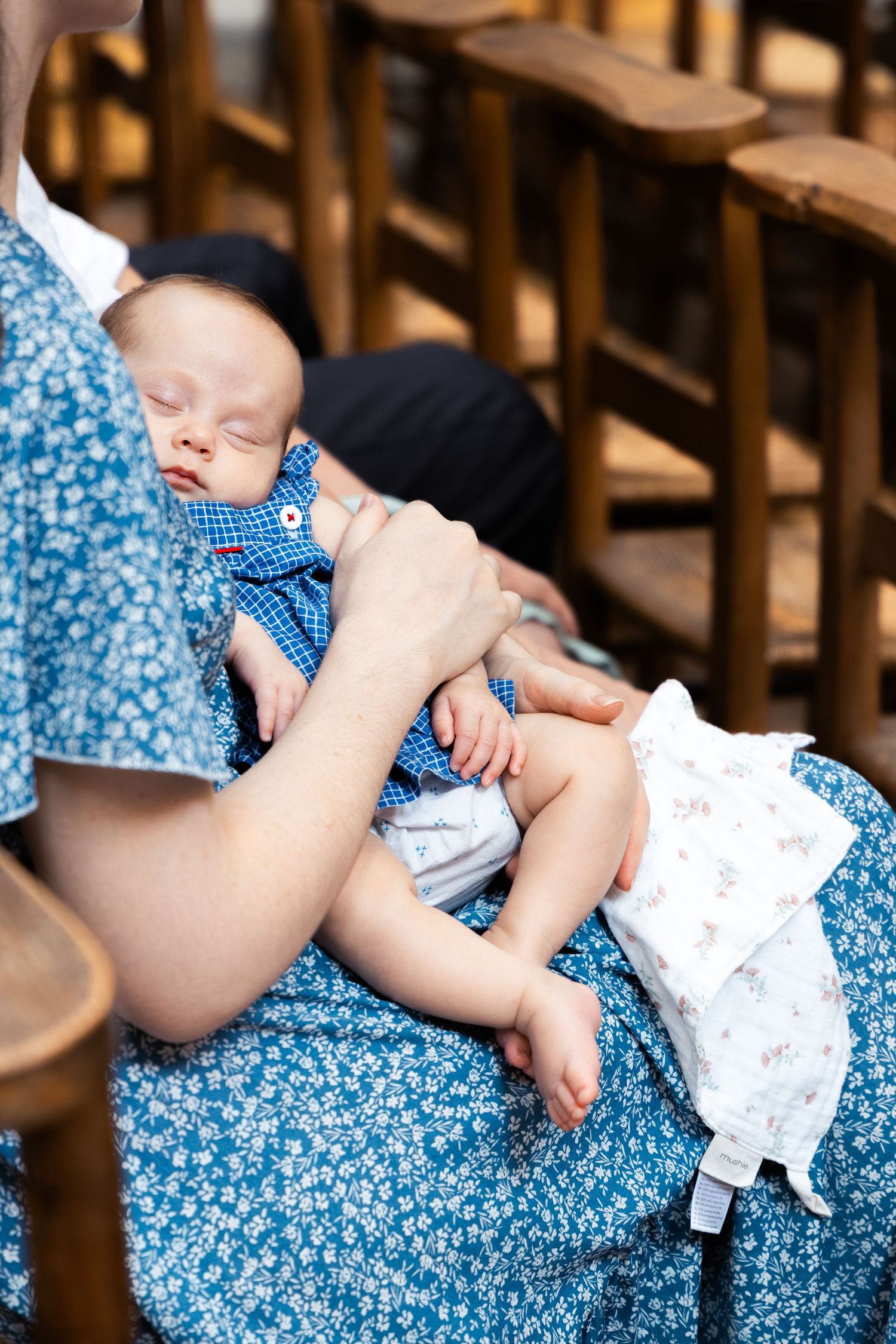 A sleeping newborn baby in blue outfit cradled in mother's arms wearing a floral blue dress, seated on wooden chairs.