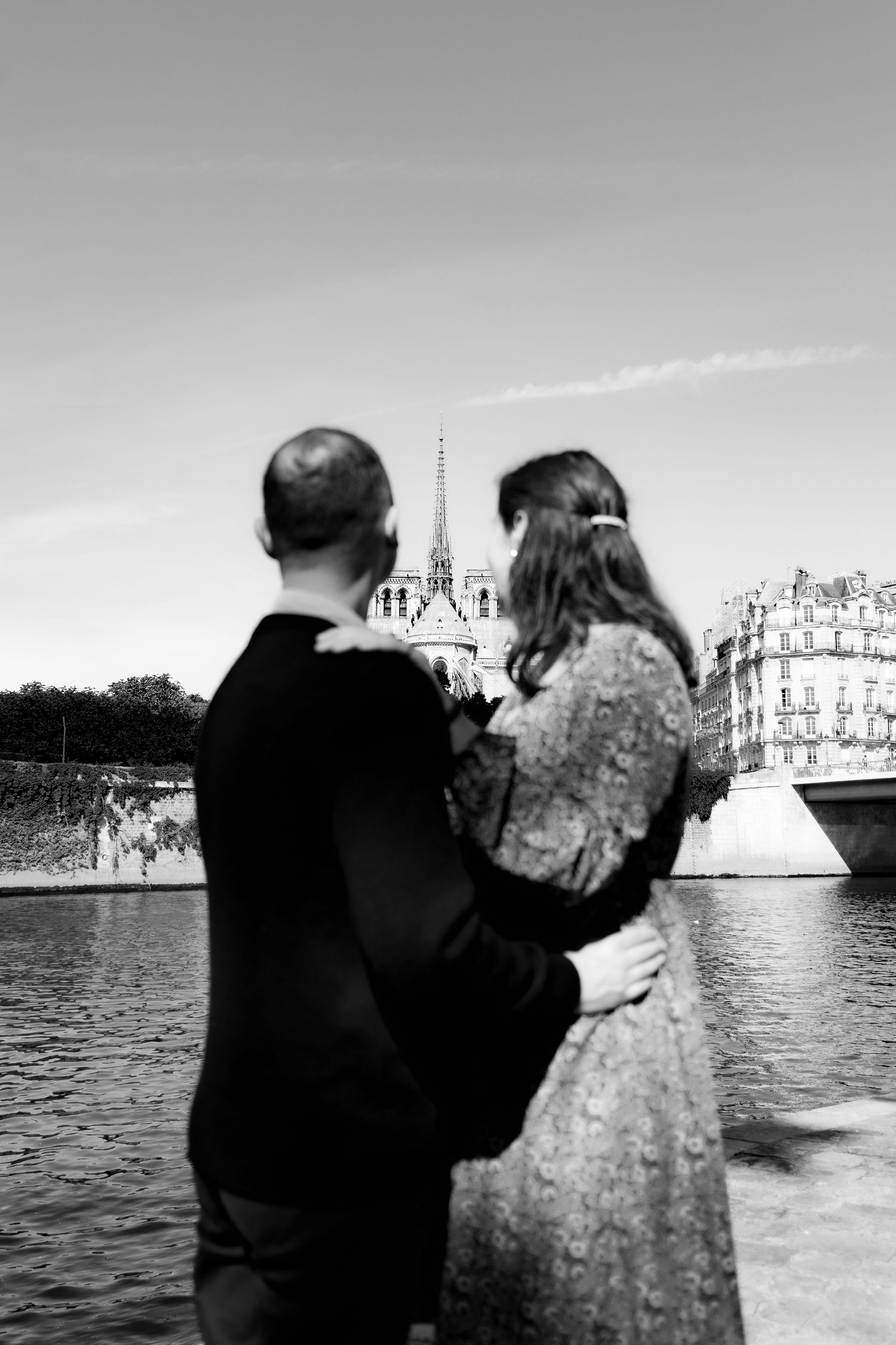 Black and white photo of a couple embracing by the Seine River with Notre-Dame Cathedral in Paris background.