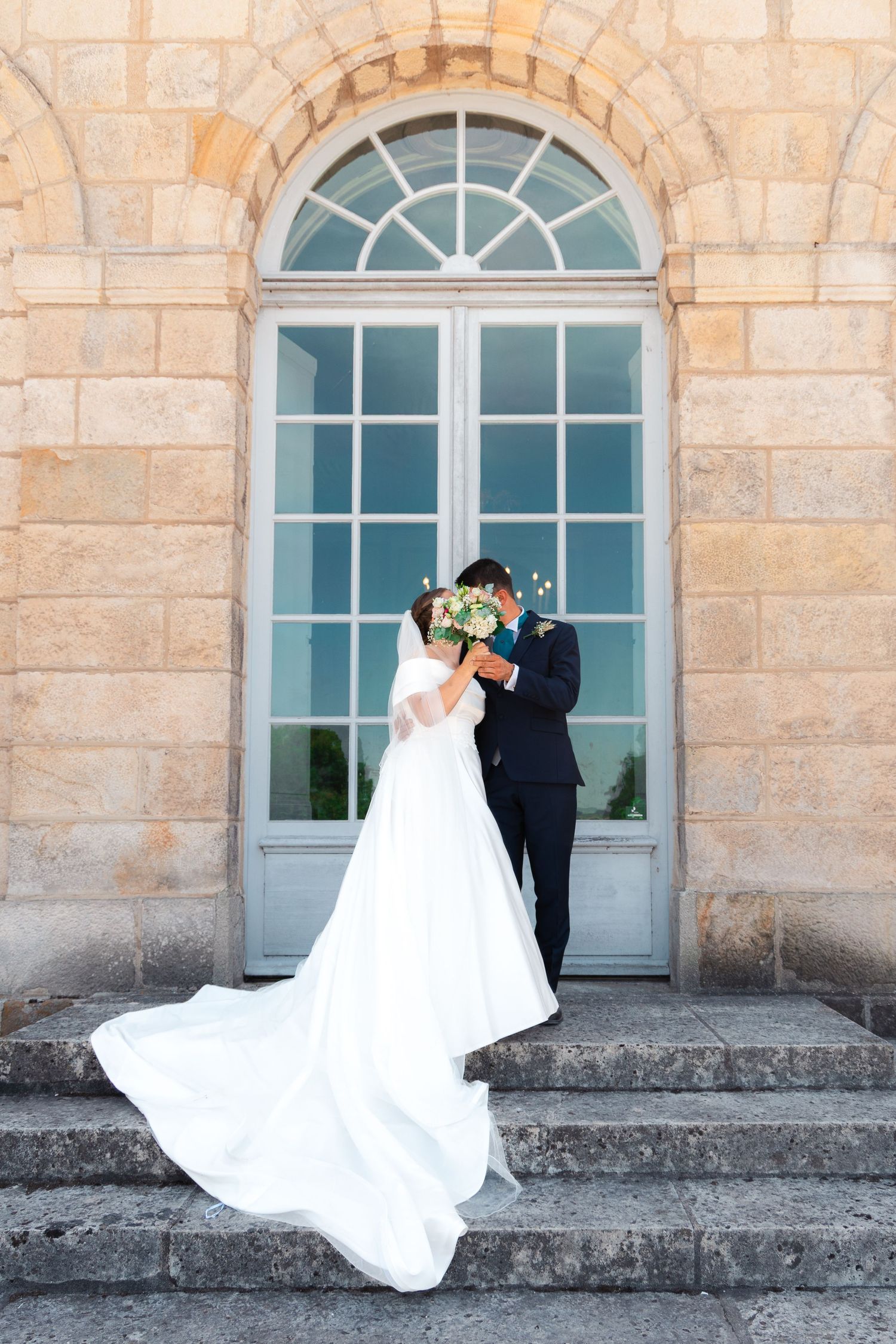 Bride and groom kissing behind bouquet on stone steps before elegant arched door in white wedding dress and dark suit.