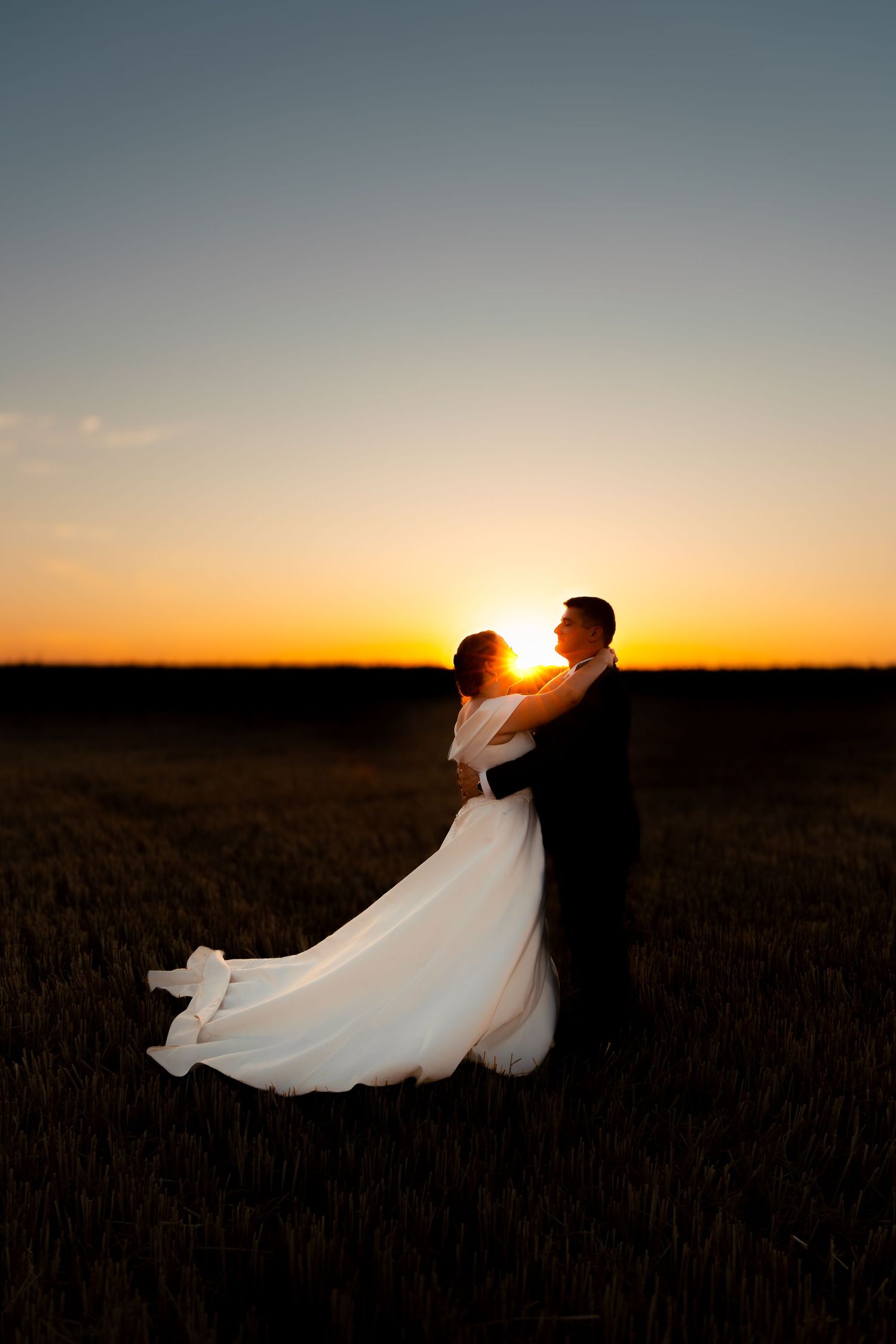 A bride and groom embrace in a wheat field at sunset, silhouetted against a vibrant orange sky.