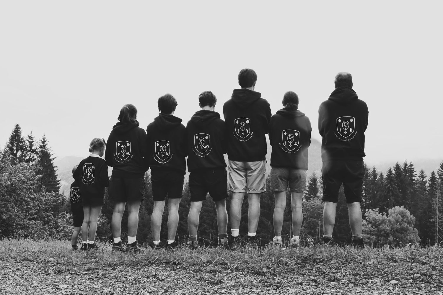 Group of seven people in matching black hoodies standing in a row overlooking a forested landscape, black and white photo.