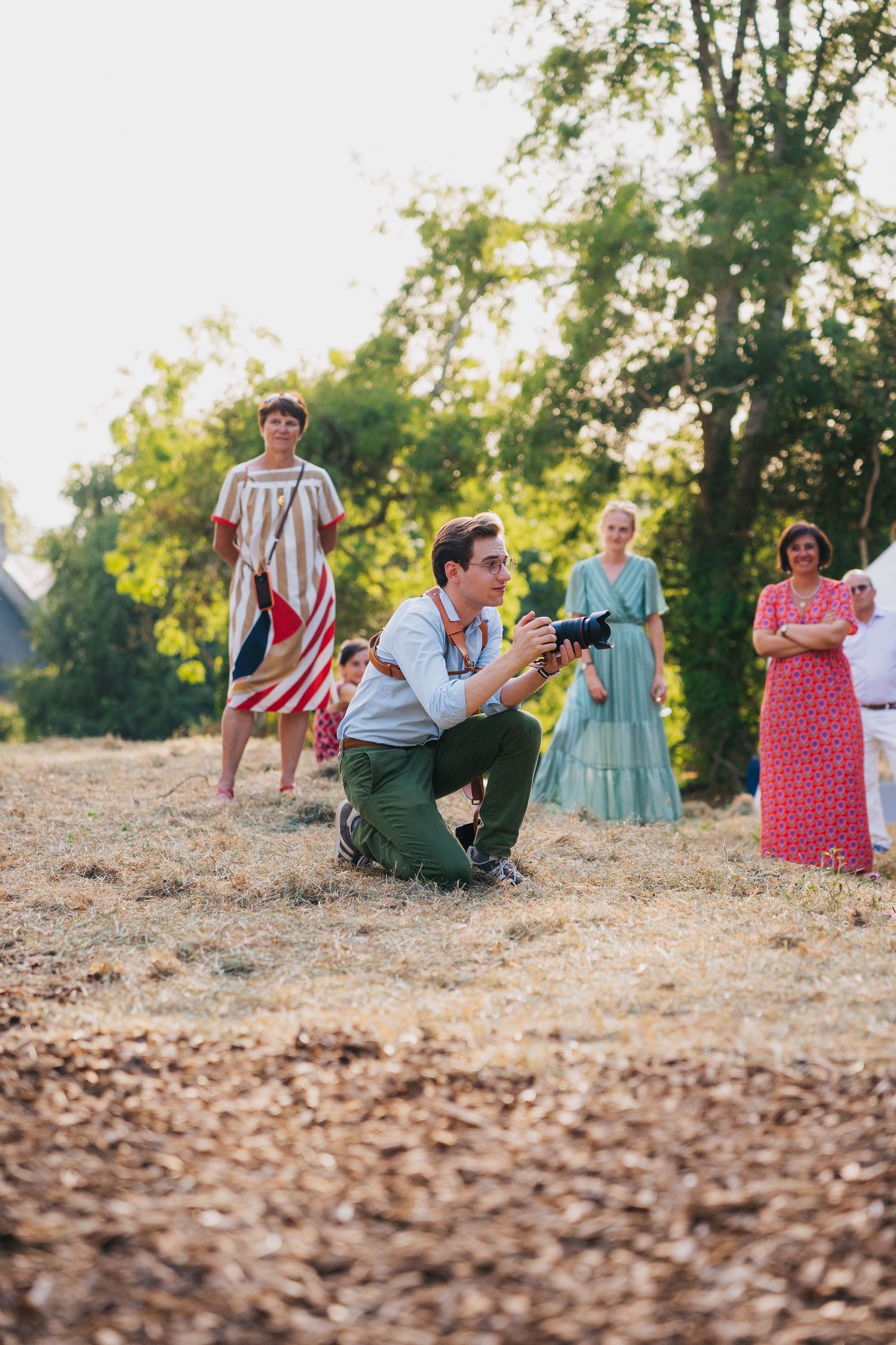 Photographer kneeling on dry grass capturing shots at an outdoor gathering with people in colorful summer attire.