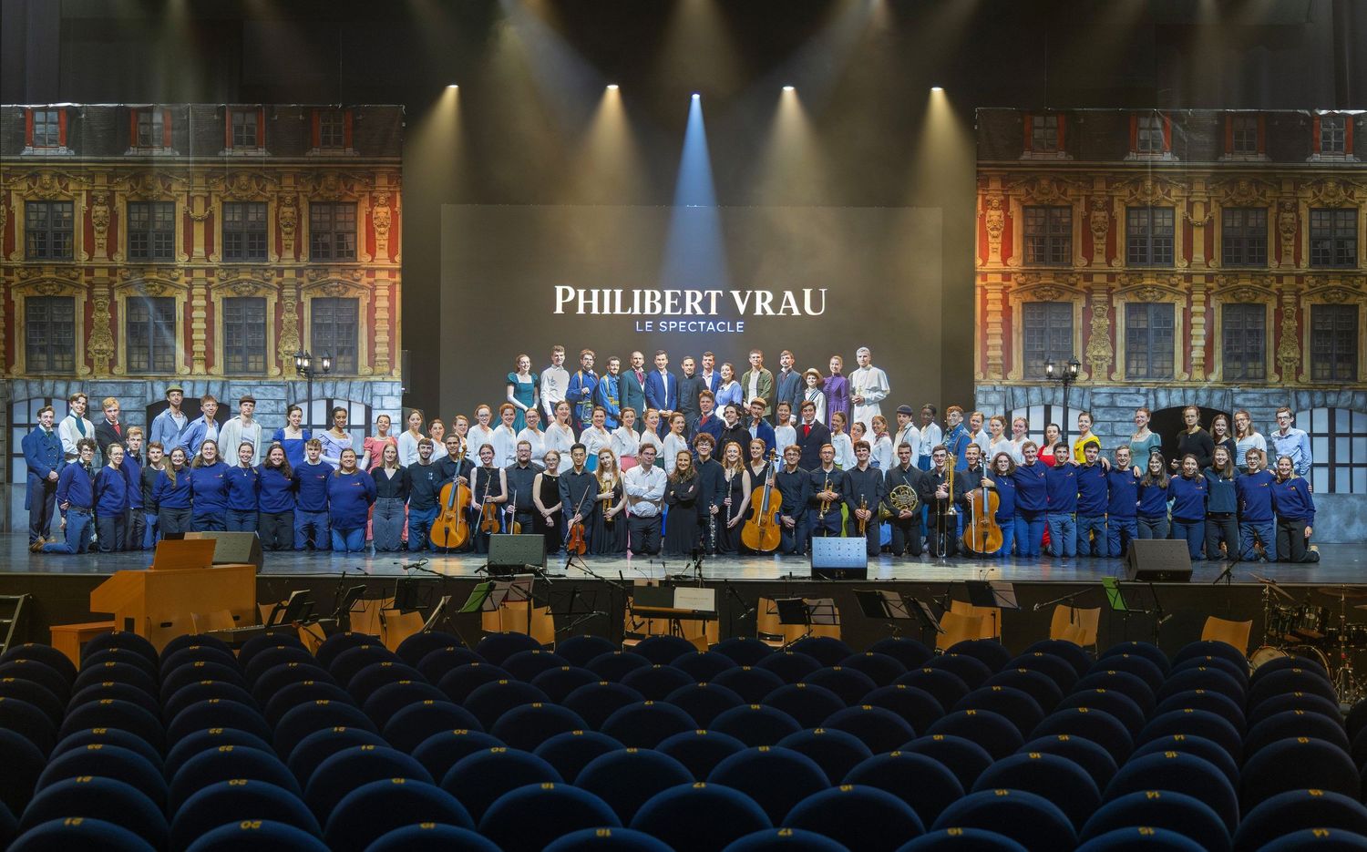 Large orchestra ensemble poses on stage under Philibert Vrau sign in an elegant concert hall.
