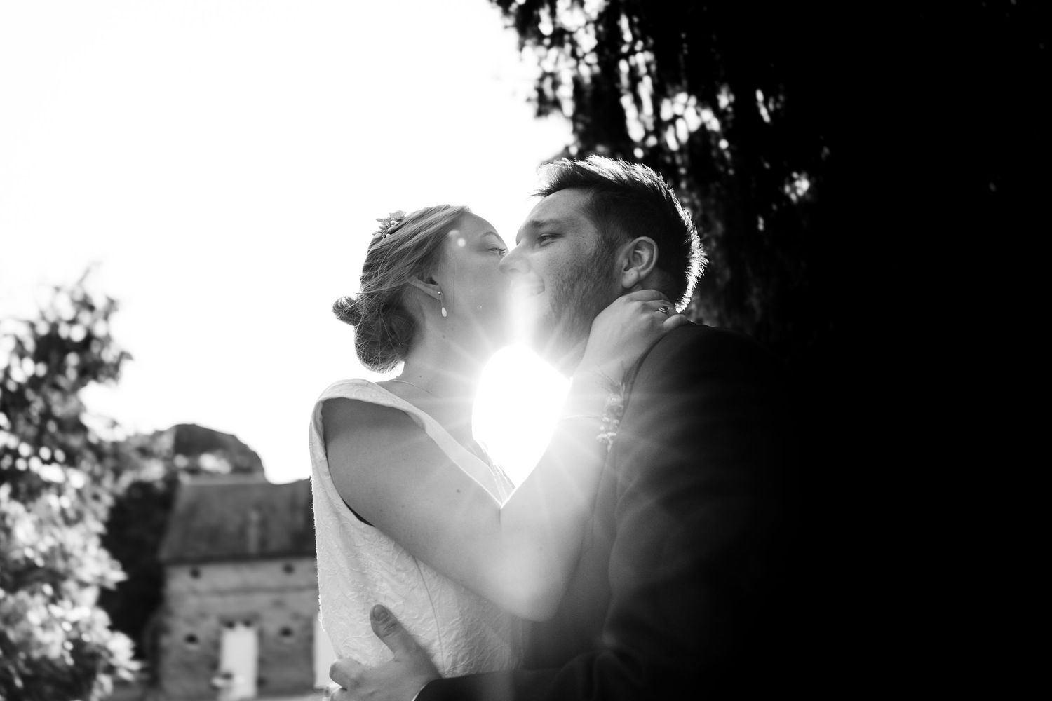 Black and white wedding photo of couple sharing a romantic kiss with sunlight glowing between them outdoors.