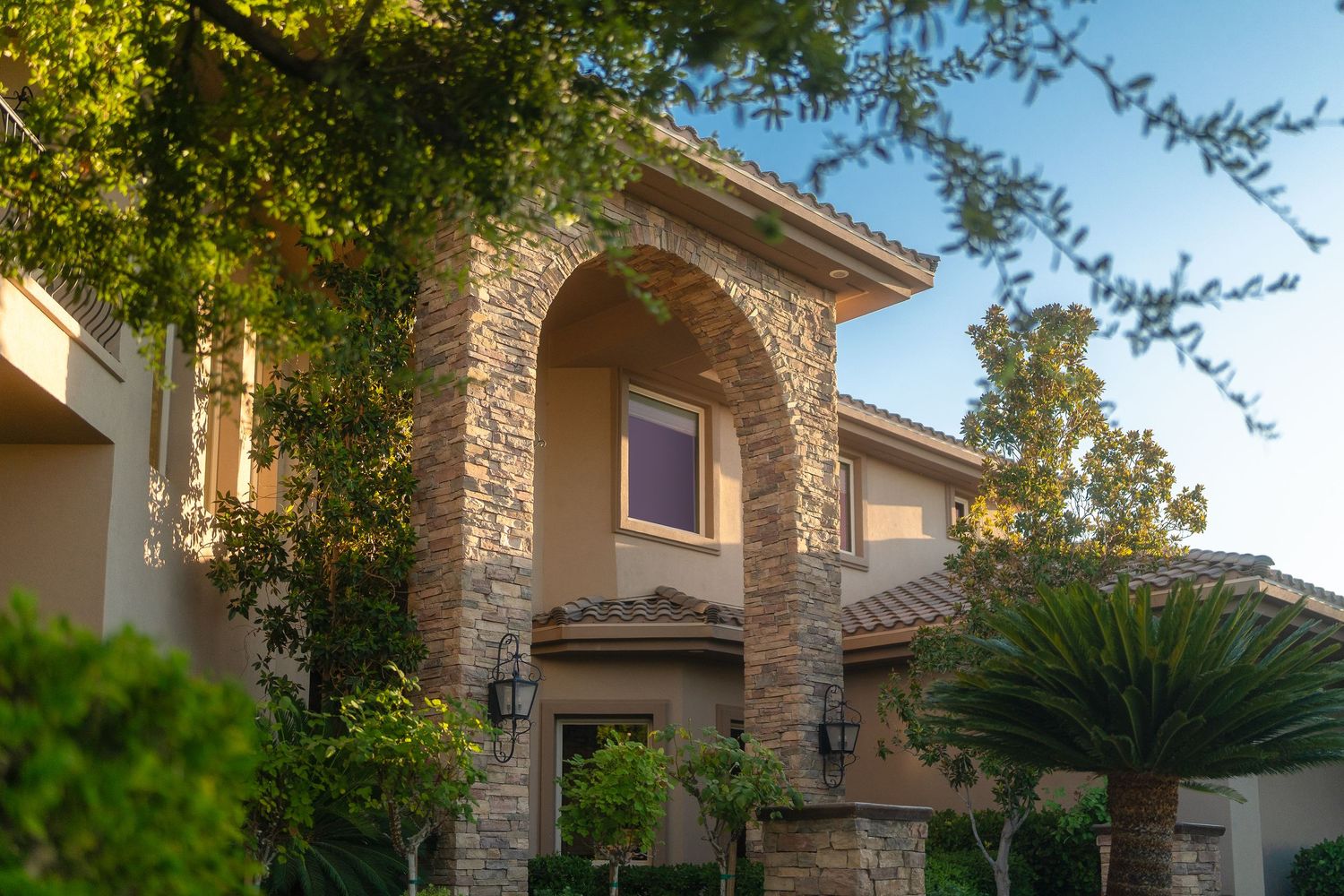 Luxury two-story home with stone archway, wall sconces, and lush green trees under a clear blue sky.