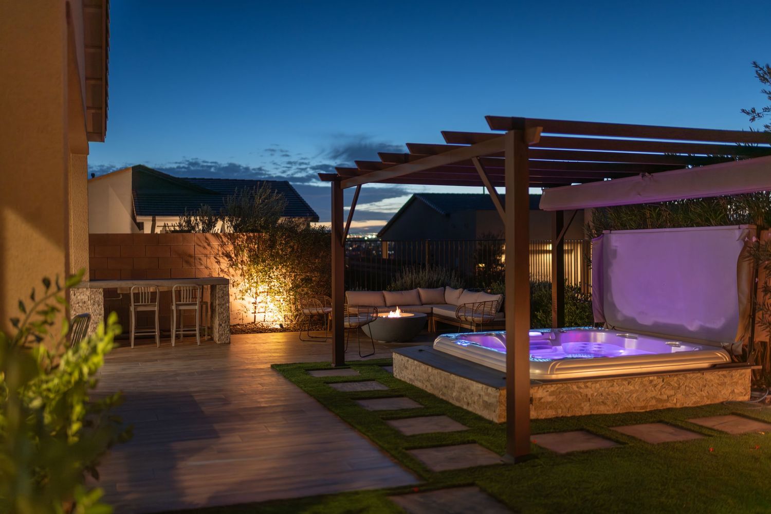 Luxurious outdoor patio with illuminated hot tub under wooden pergola at dusk, featuring lush garden and ambient lighting.