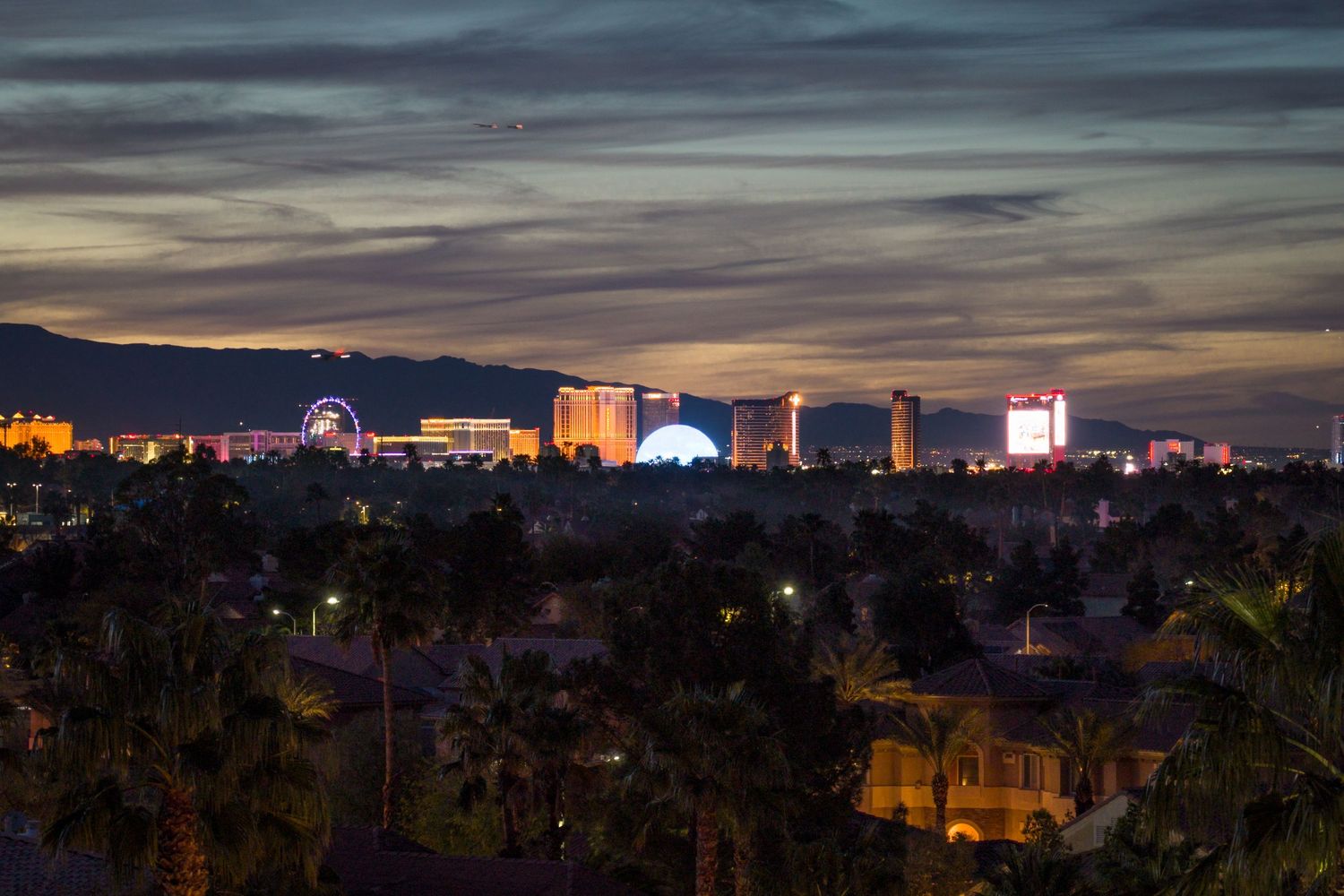Las Vegas Strip skyline at dusk with colorful casino lights, Ferris wheel, and palm trees silhouetted against a dramatic sky.