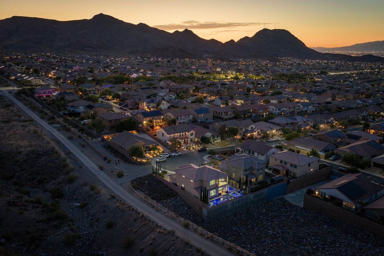 Aerial dusk view of a suburban neighborhood with glowing homes, mountain backdrop, and colorful sunset sky in a desert city.