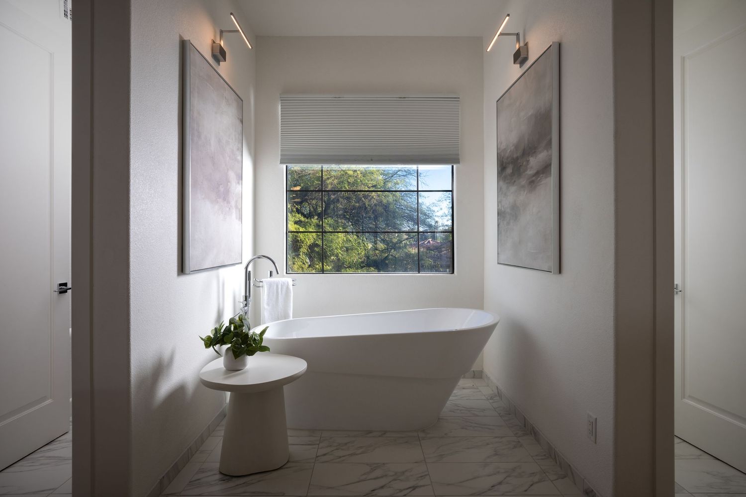 Elegant freestanding soaking tub centered beneath a window, flanked by abstract art and marble floors in a modern bathroom.