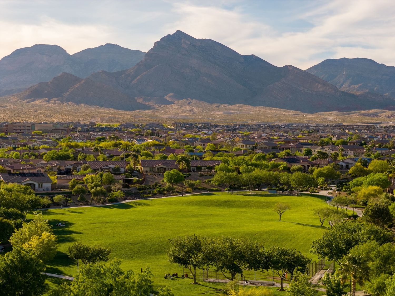 Aerial view of a lush green golf course with desert homes and stunning mountain backdrop under a partly cloudy sky.