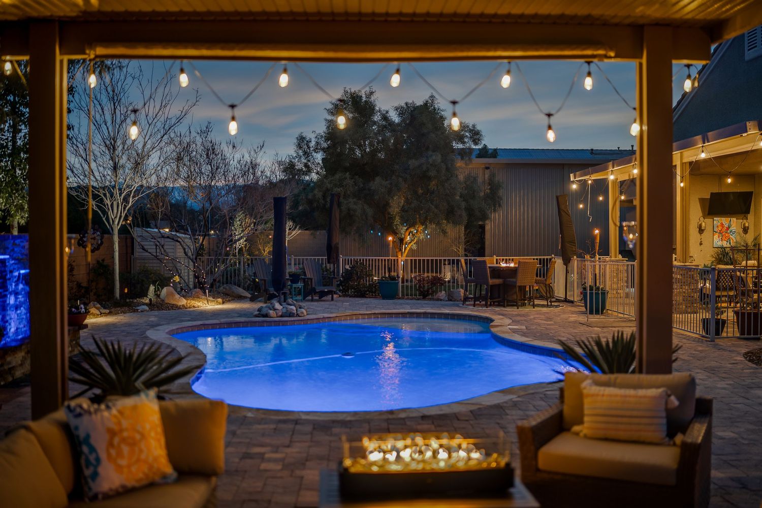 Luxurious backyard pool area illuminated by string lights with cozy seating and fire features at dusk.