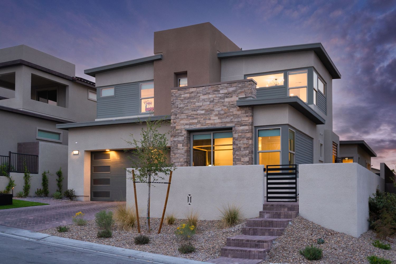 Modern two-story desert home with stone accents, flat roof, and warm interior lighting at dusk in a residential neighborhood.