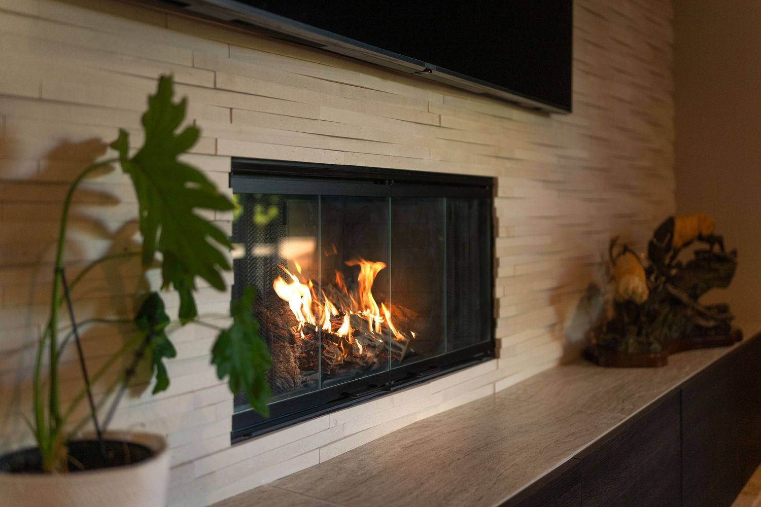 Modern linear gas fireplace with glowing flames set in a light stone wall with a TV above and green plants nearby.