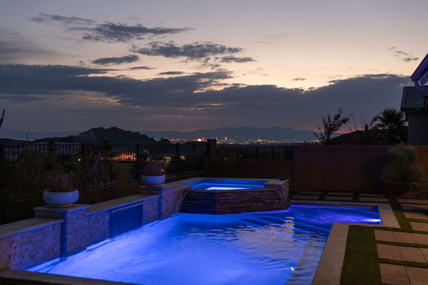 Illuminated luxury pool and spa glowing blue at dusk with city lights and mountains in the background.
