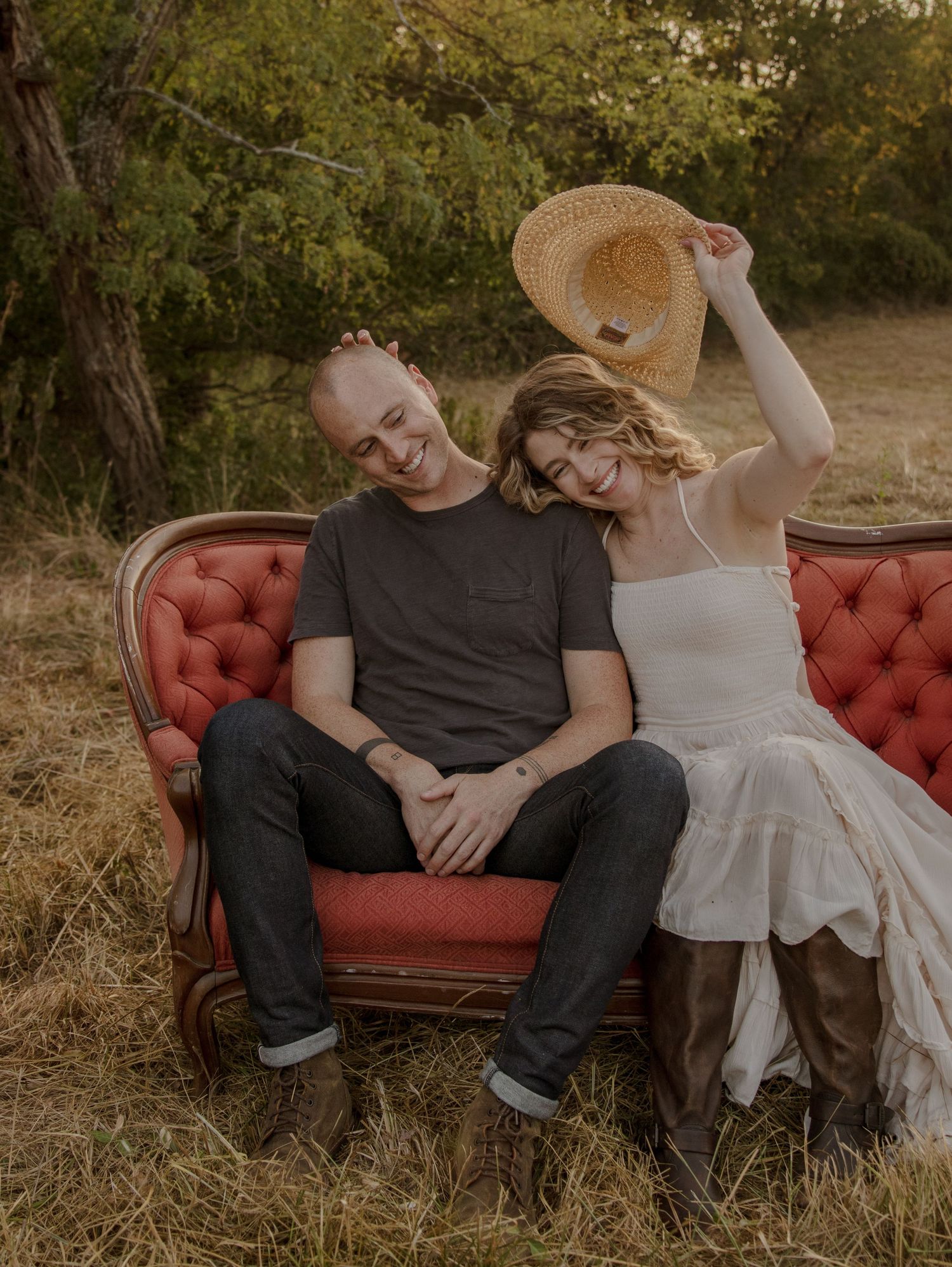 A couple sits together on a vintage red velvet couch outdoors during a warm sunset photo session in the woods.