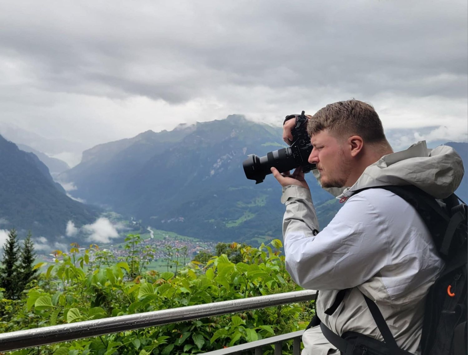 Photographer with backpack capturing mountain valley landscape with camera on cloudy day at scenic overlook.