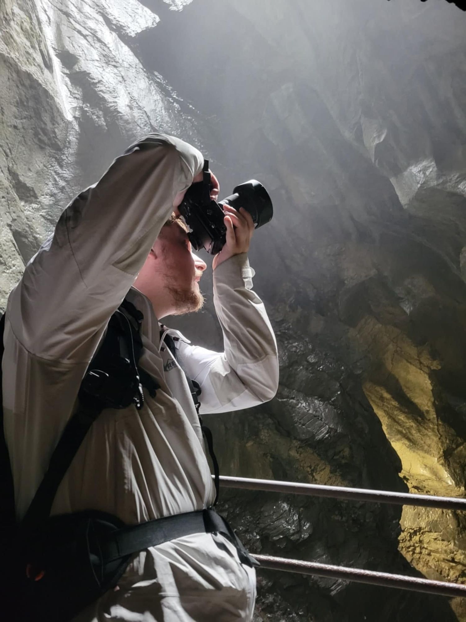 A photographer in a hooded jacket captures images inside a dramatic sunlit cave.