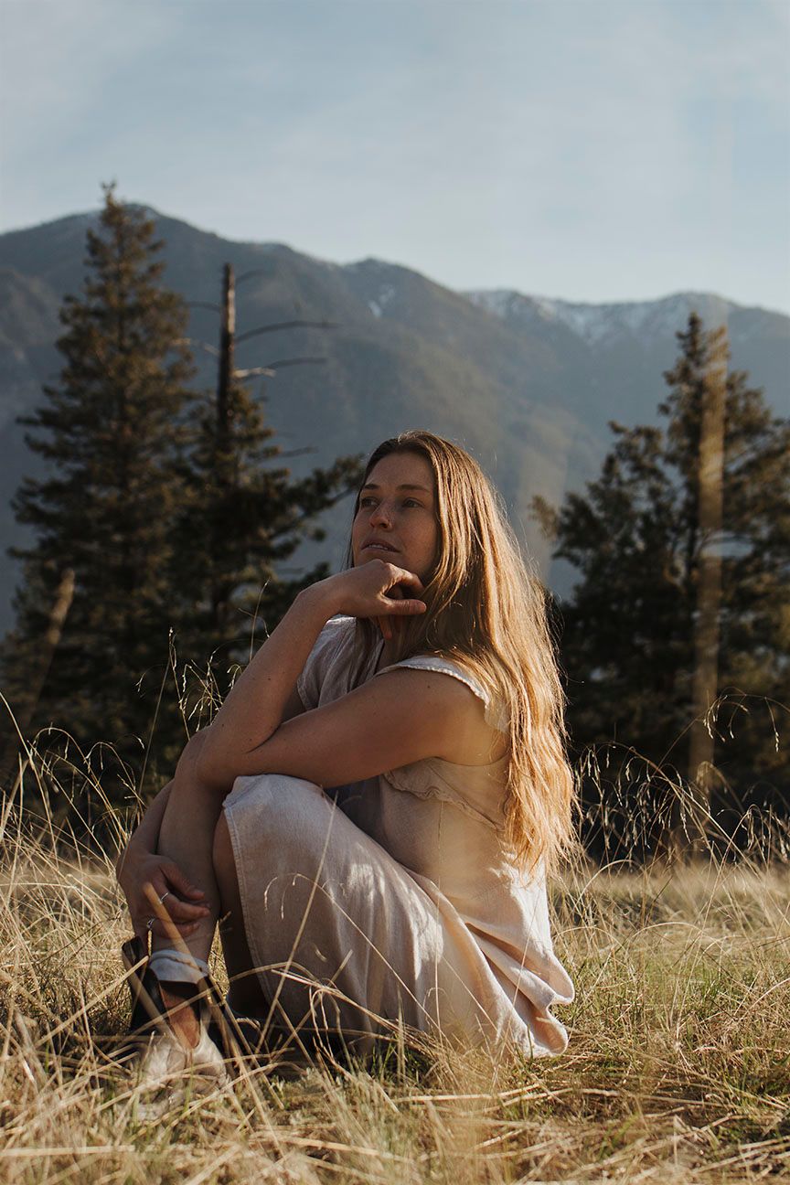 Someone in casual attire sits contemplatively on a grassy hillside with mountain peaks and evergreen trees in the background.