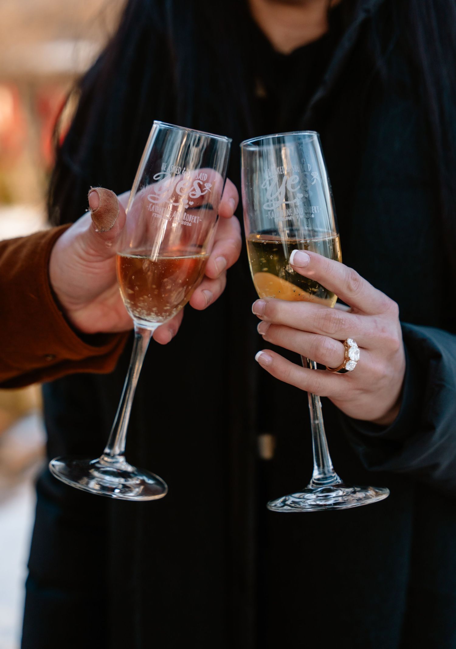Two people toasting with engraved champagne flutes, one wearing an engagement ring, celebrating outdoors.