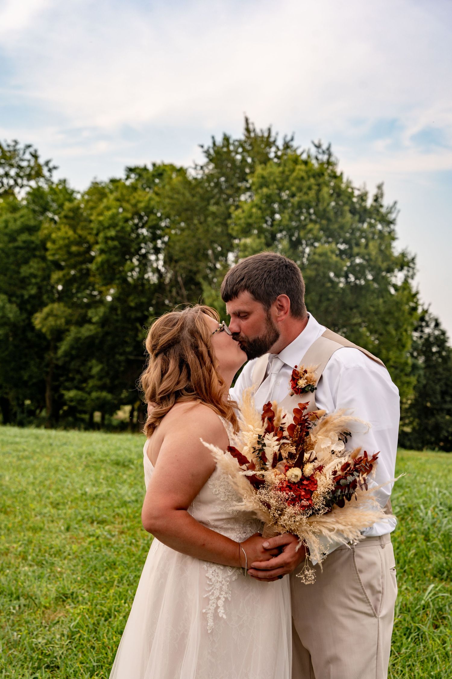 A bride and groom share a kiss in a green field, she holds a rustic dried floral bouquet in warm autumn tones.