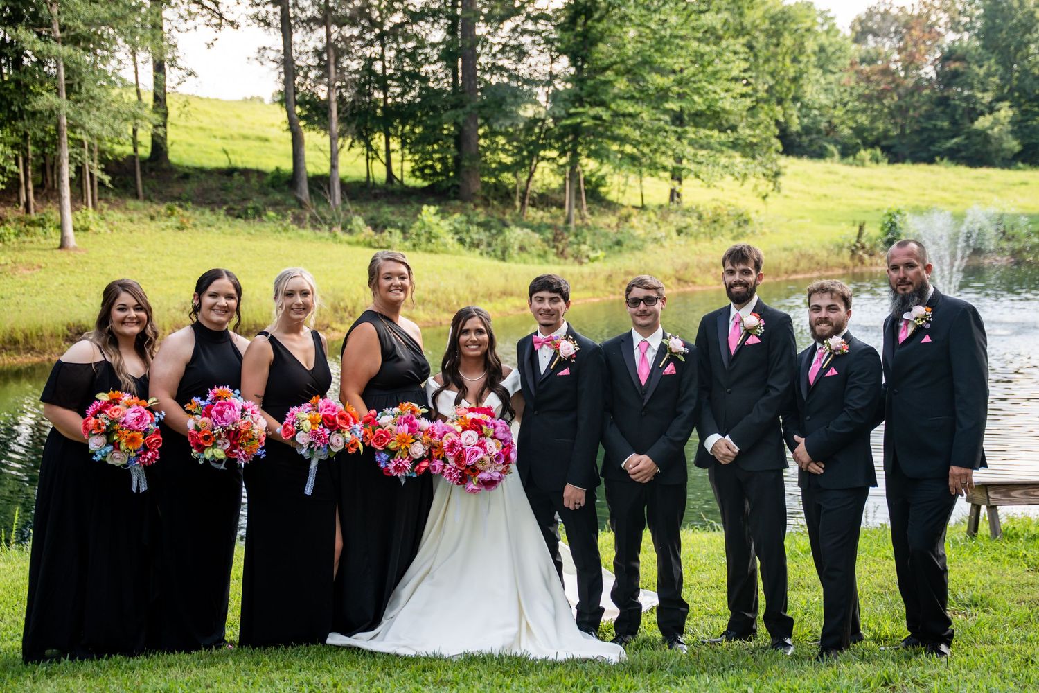Wedding party posing outdoors, bridesmaids in black gowns with colorful bouquets, groomsmen in suits with pink ties, scenic pond backdrop.