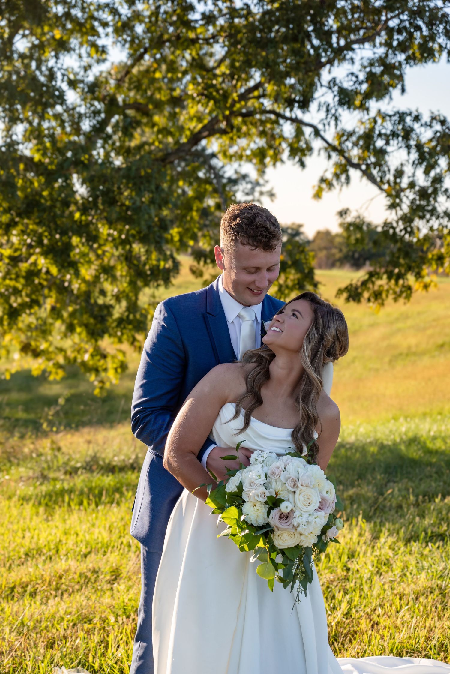 Bride and groom embrace outdoors in golden hour light, she holds white bouquet, he wears navy suit, green trees behind them in Chaplin, Ky
