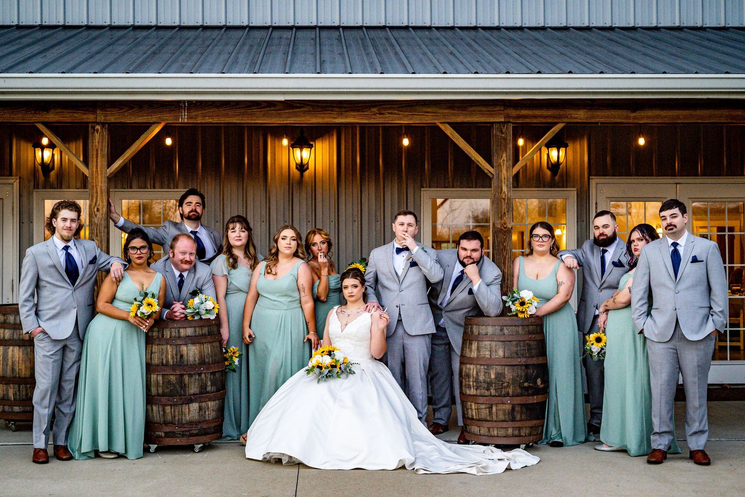 Wedding party poses with bride and groom beside rustic barrels outside a barn venue, bridesmaids in sage green gowns.
