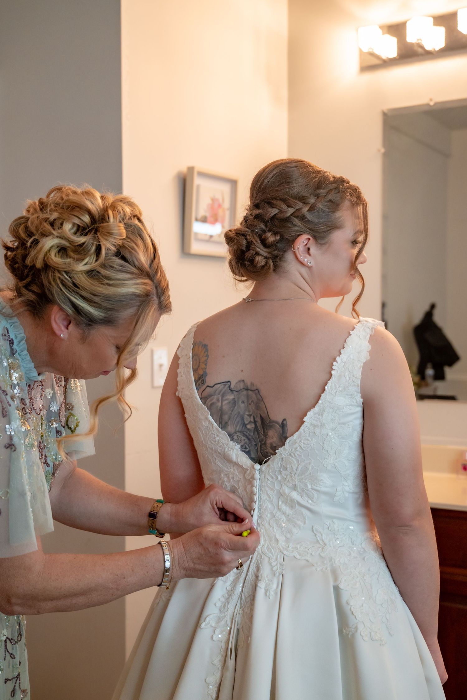 Woman helping bride button up lace wedding dress, both with elegant updo hairstyles, bride showing back tattoo.
