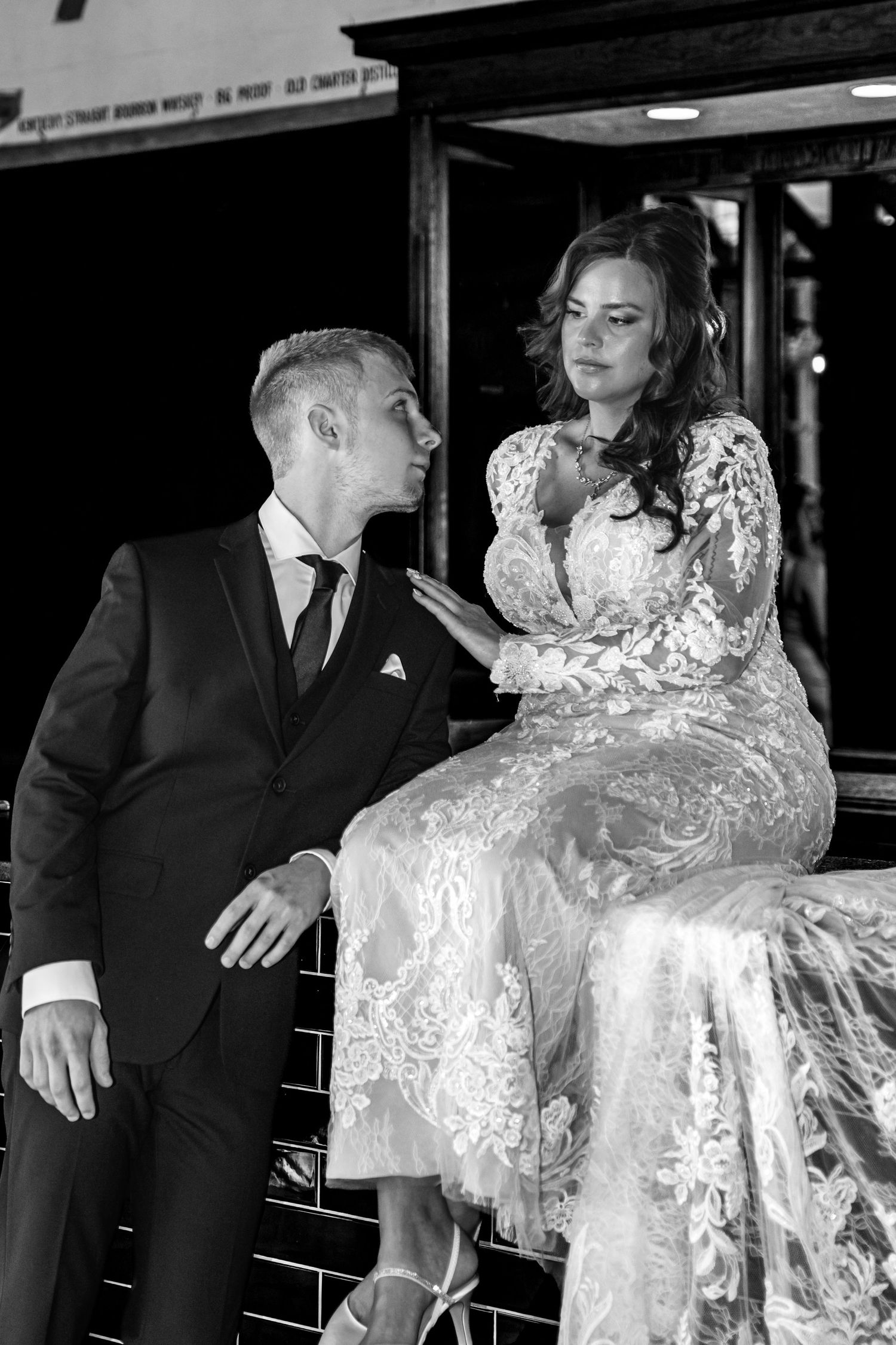 Black and white wedding photo of groom gazing up at bride in lace dress seated on railing.