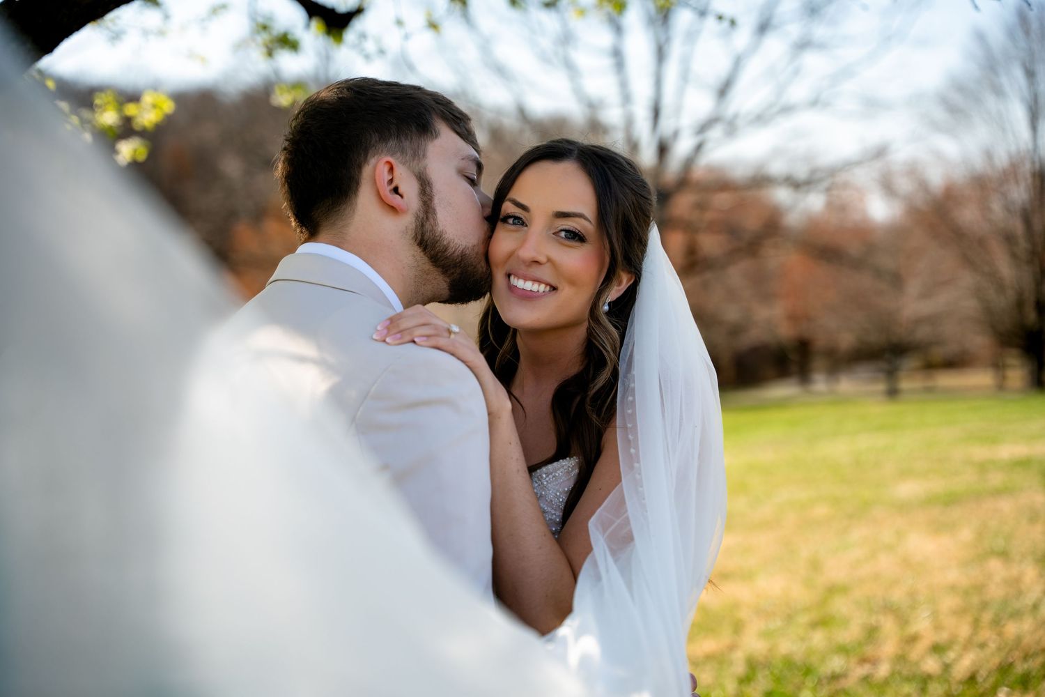 Groom kissing bride on cheek outdoors, bride smiling in white gown and veil on sunny wedding day.