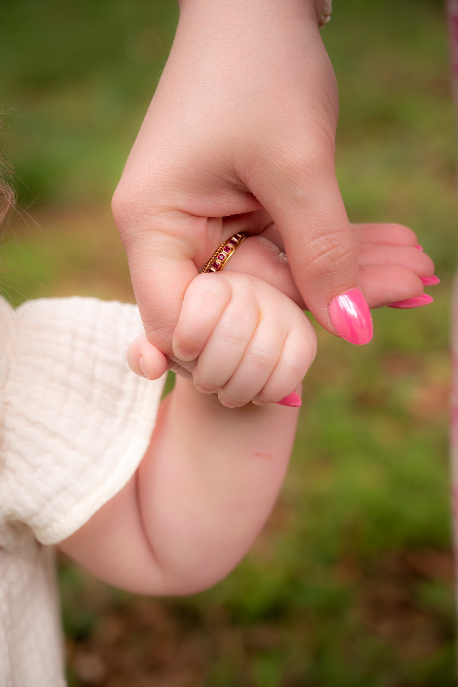A mother with pink nails and a gold ring holds her small child's tiny hand outdoors.