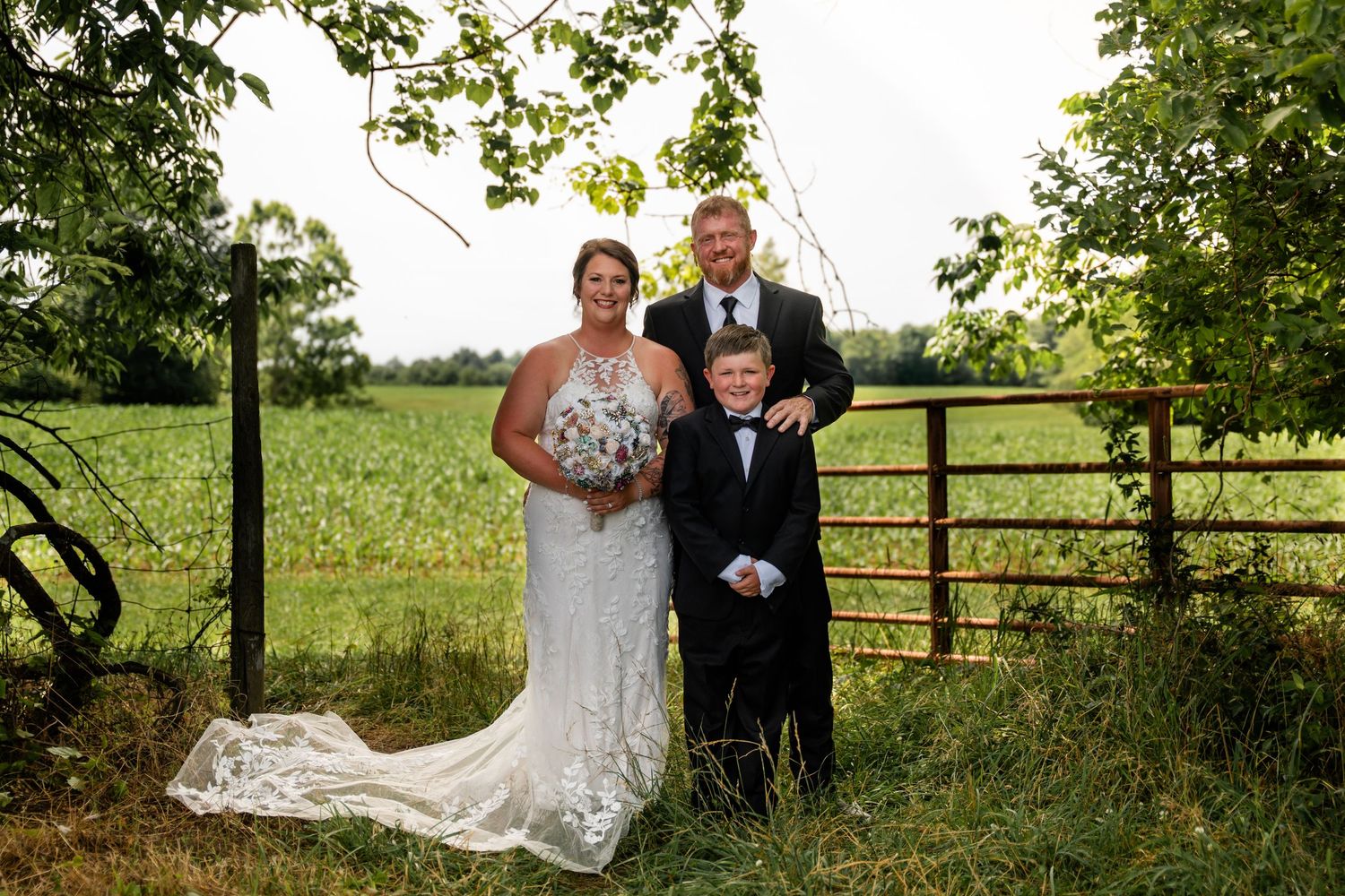Wedding couple with young boy posing by rustic farm gate in lush green countryside setting.