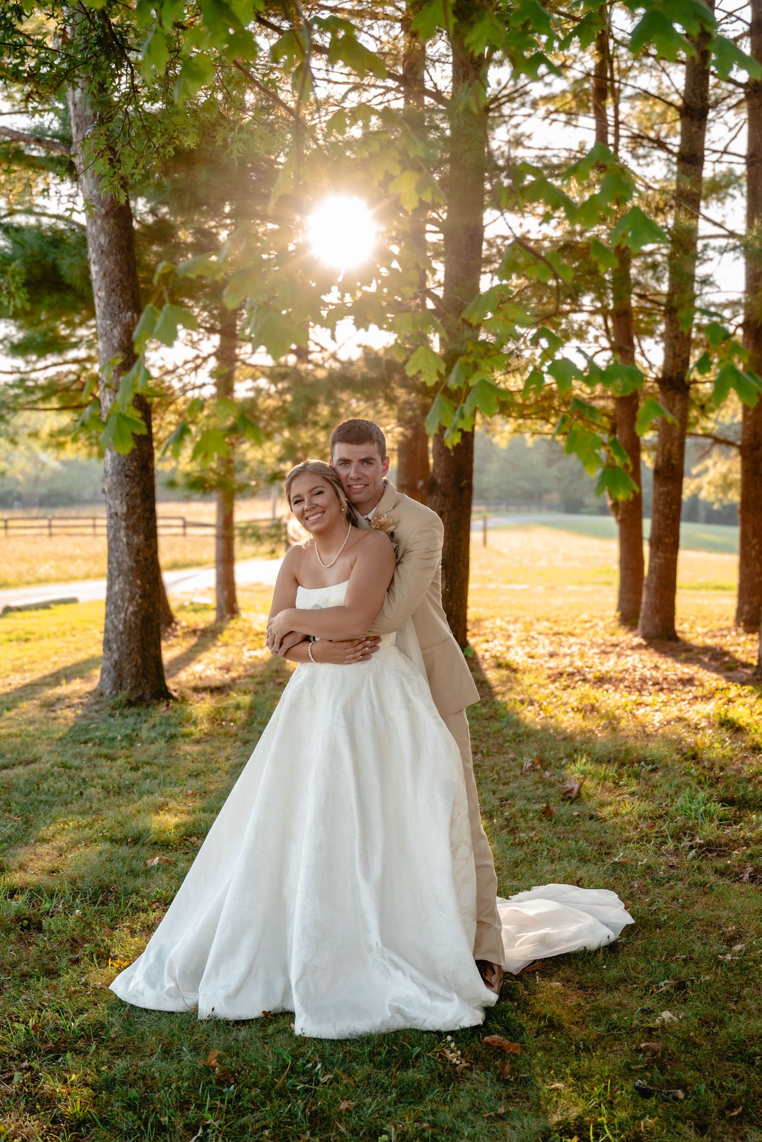 Bride and groom embracing at golden hour sunset in a lush green tree-lined outdoor wedding venue.
