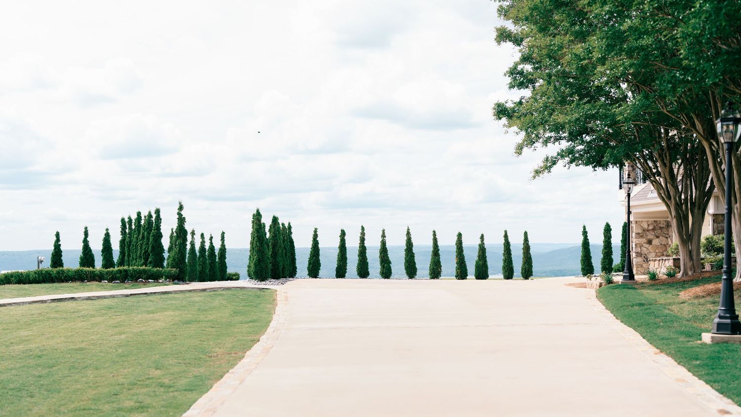 A serene driveway lined with tall cypress trees leads to an open landscape under a cloudy sky.