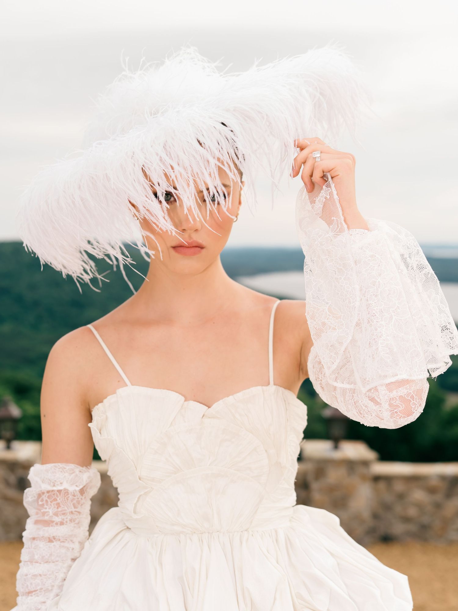 Bride poses with white feather fascinator and lace gloves in strapless gown with scenic backdrop.