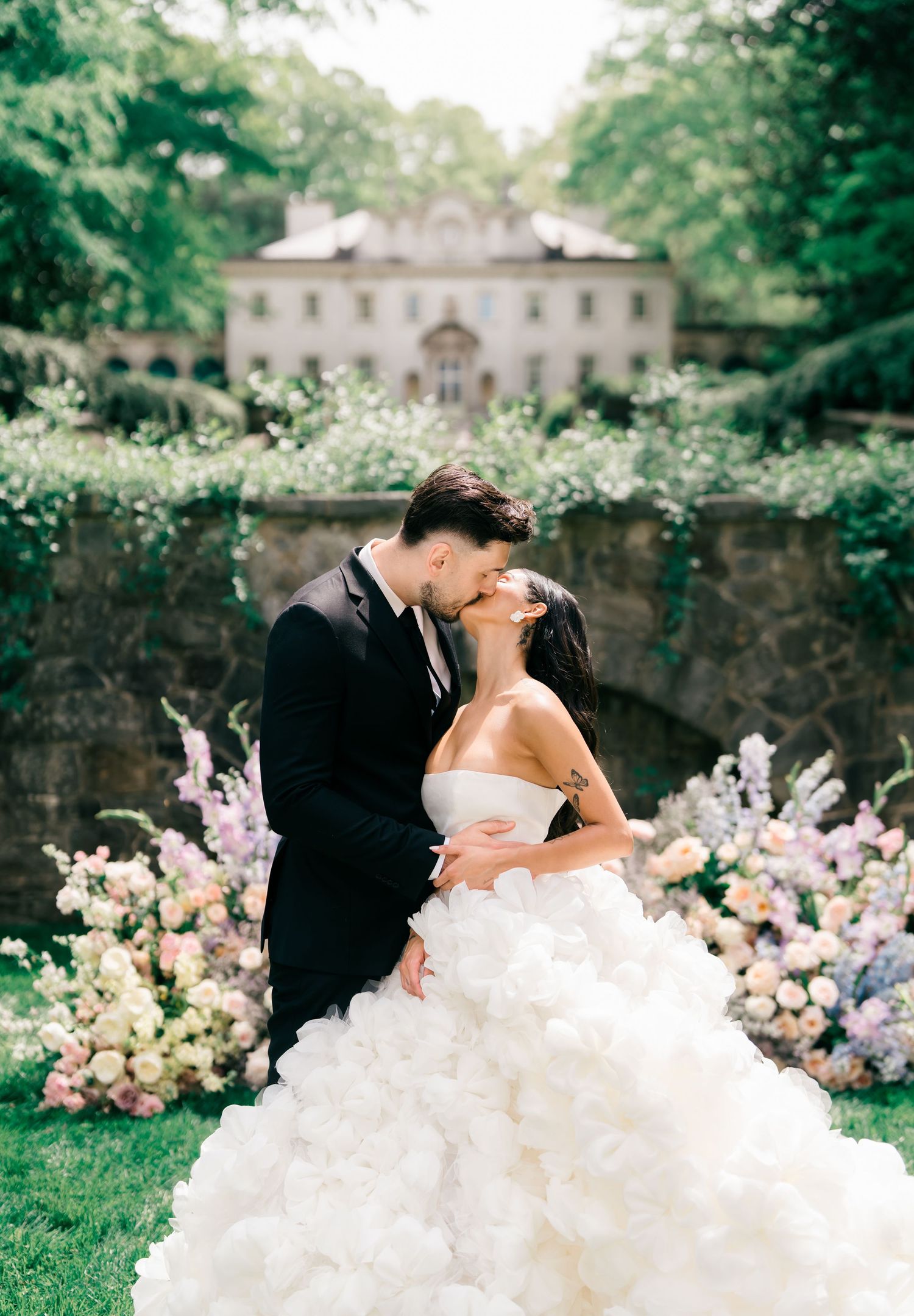 Bride and groom share a romantic kiss in a stunning garden wedding setting with lush floral arrangements and an elegant estate backdrop.