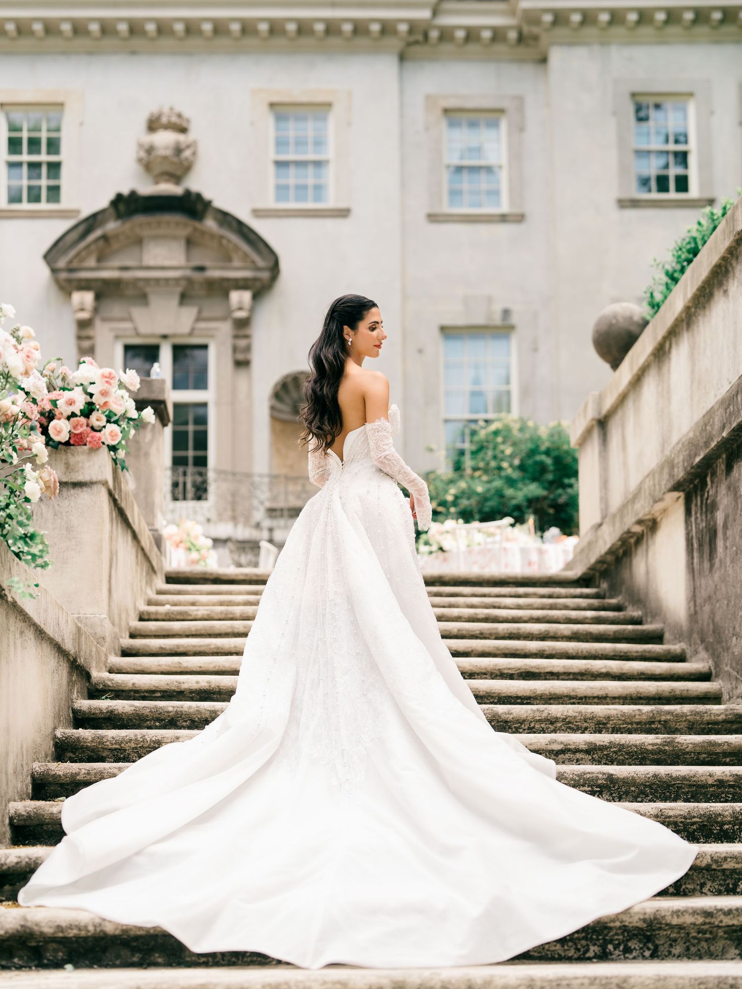 Bride in elegant white ball gown with long train stands on stone steps of grand manor house.