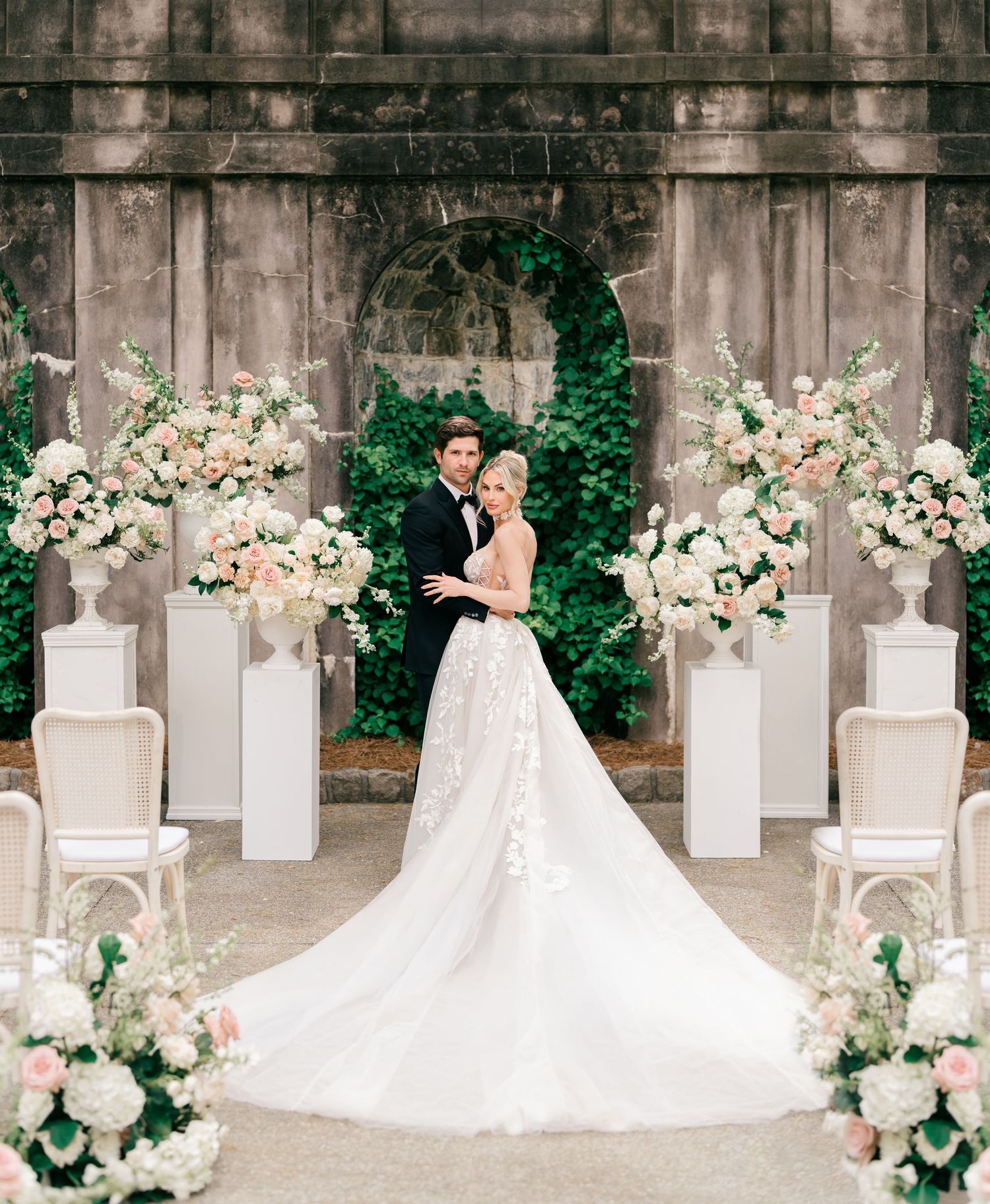 Elegant wedding couple posing at floral ceremony altar with white and pink blooms, lush greenery backdrop, and bride in lace gown.