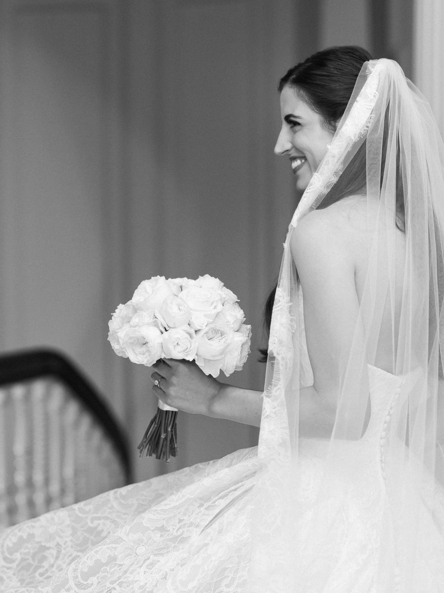 Smiling bride in lace gown and veil holds white rose bouquet in elegant black and white wedding portrait.