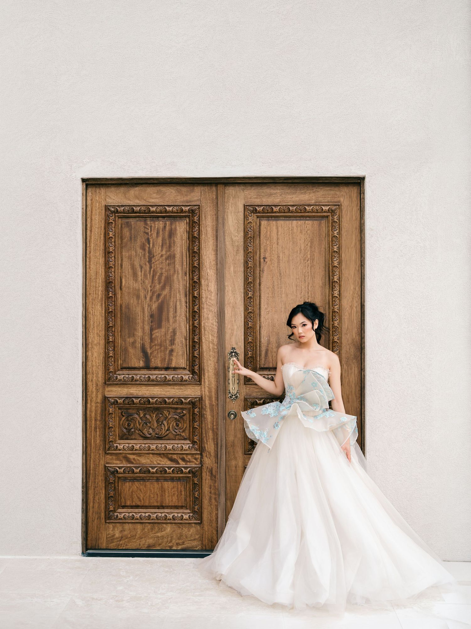 Elegant bride in strapless ballgown with blue bow detail standing before ornate carved wooden double doors.