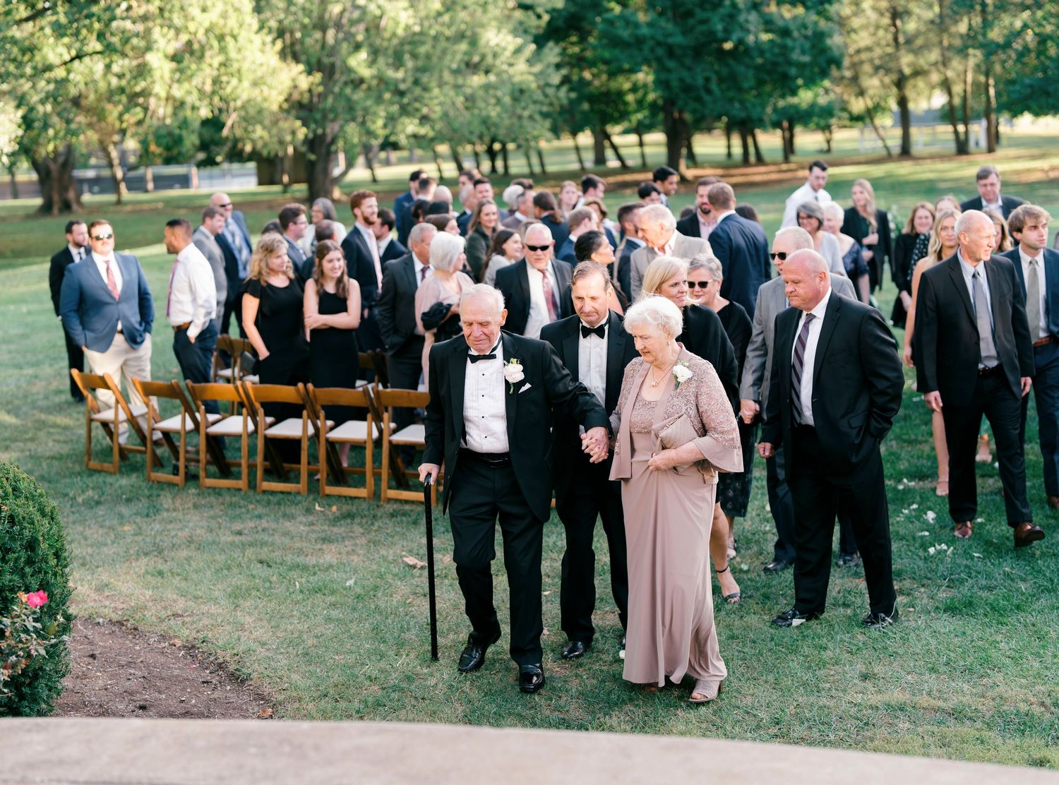 Wedding guests gather outdoors on a grassy hillside with wooden chairs arranged for an outdoor ceremony.