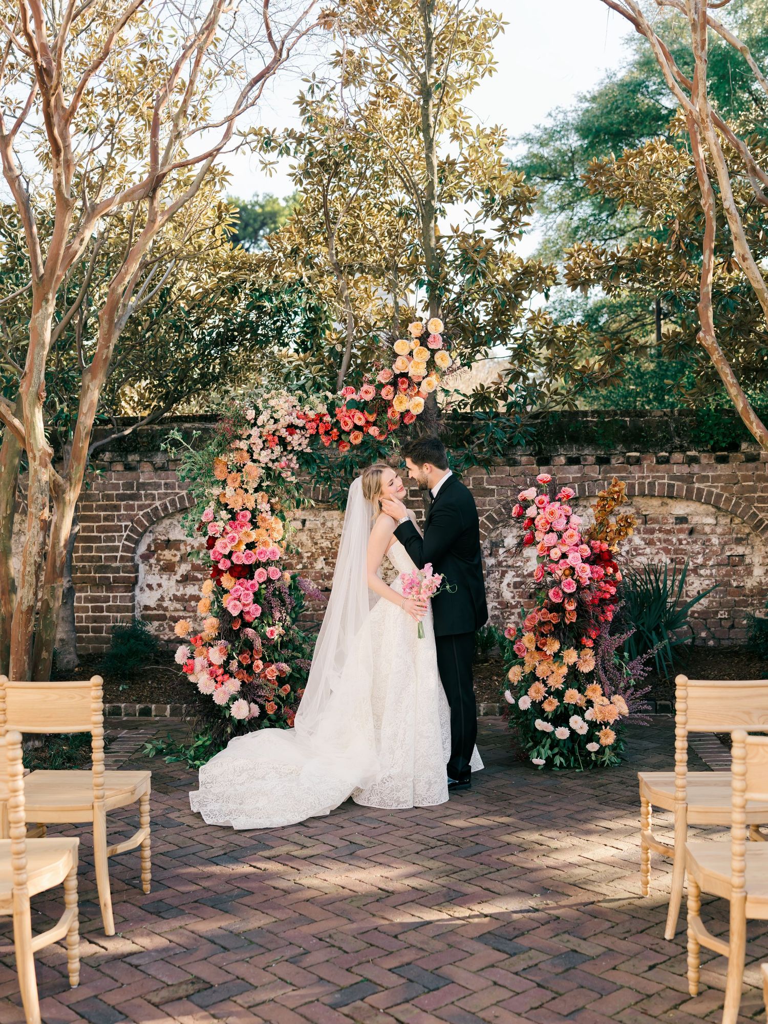 A panoramic view of an outdoor wedding ceremony setup with wooden chairs and pink floral arrangements against a brick wall.