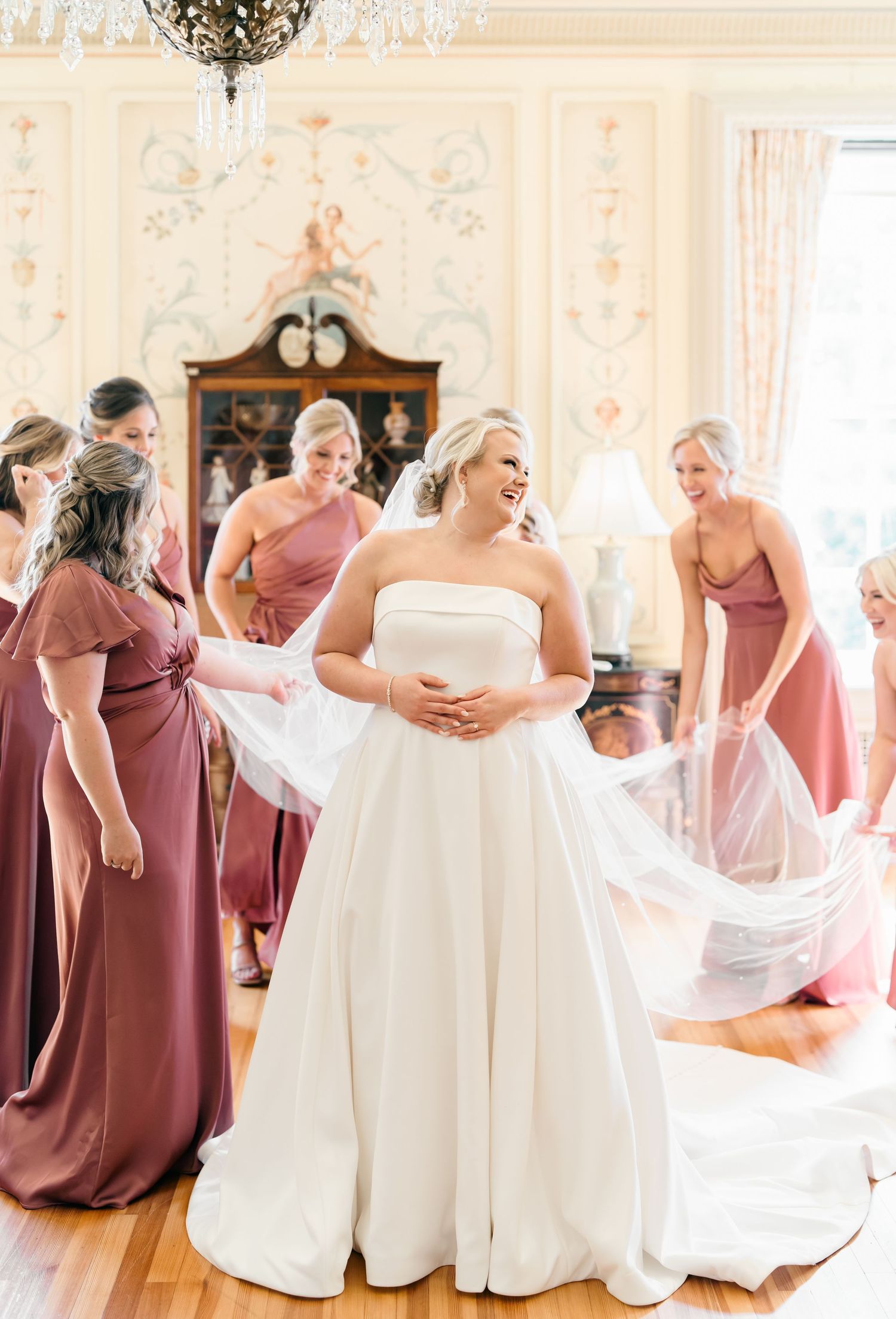 Bridesmaids in mauve dresses help the bride with her elegant white wedding gown in an ornate ballroom setting.