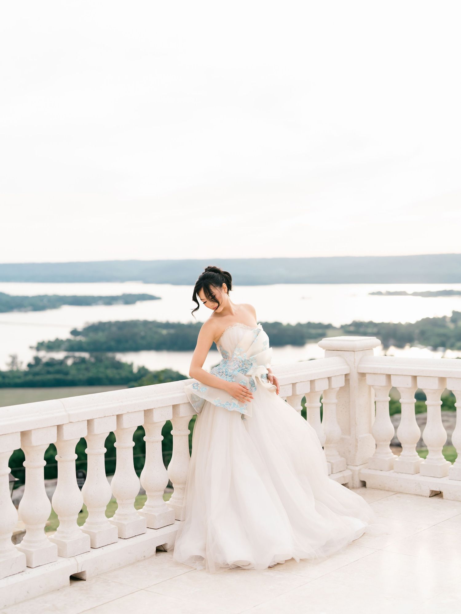 Bride in strapless ballgown leans on white balcony railing with scenic lake view behind her.