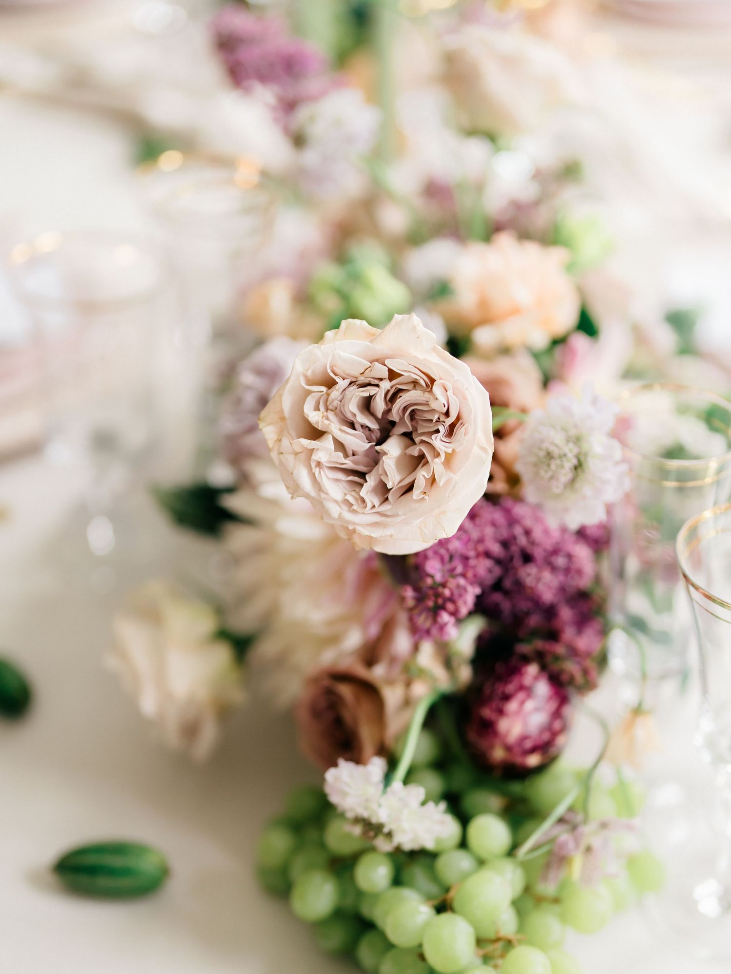 Elegant floral arrangement with blush roses, purple blooms and green accents on a light background.
