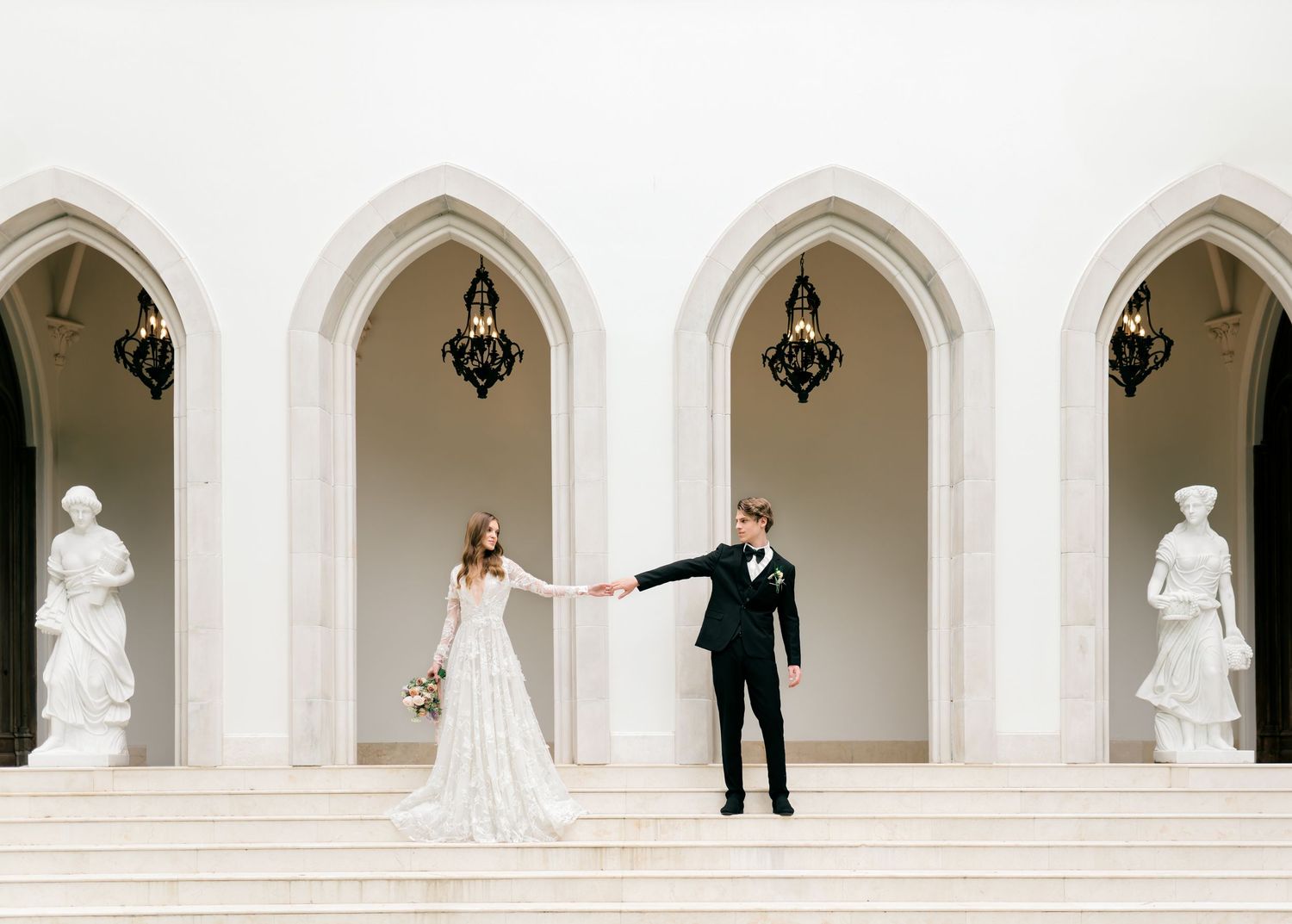 A panoramic wedding photo showing a bride and groom with their wedding party positioned in white arched hallways.