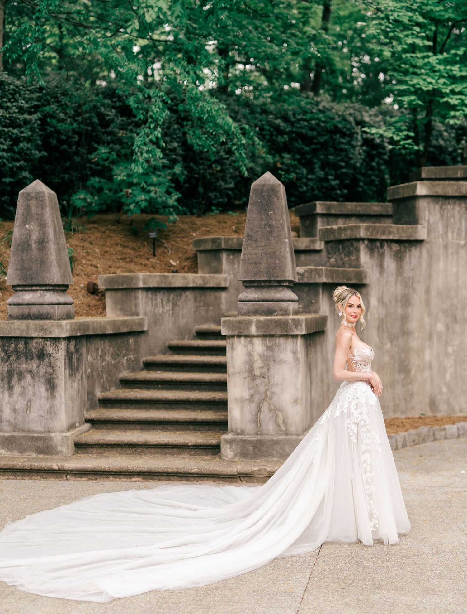 Bride in a flowing white gown with long train poses gracefully by stone steps and obelisks in a lush garden setting.