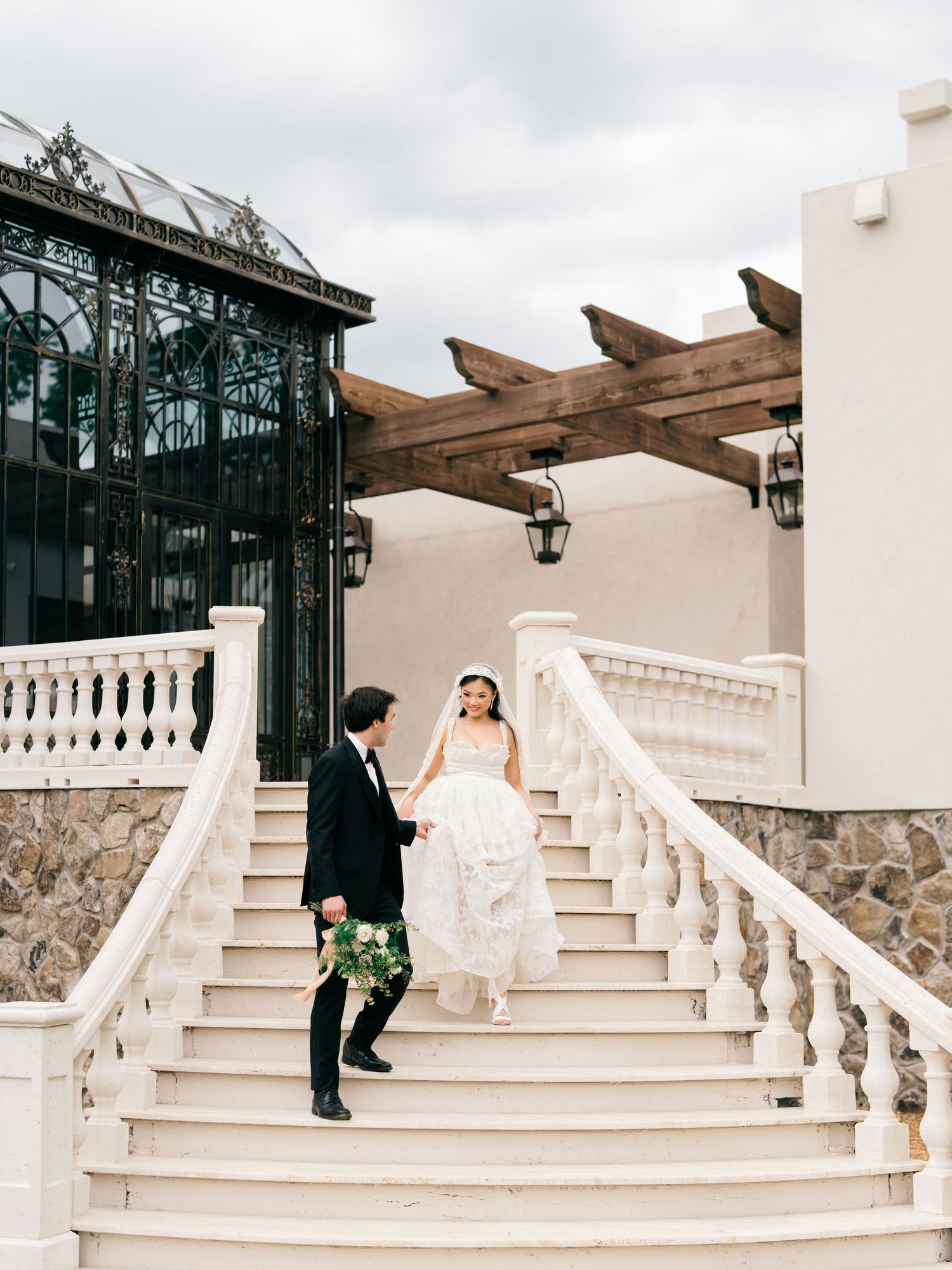 Bride and groom descend grand white staircase with ornate balustrade at elegant wedding venue.